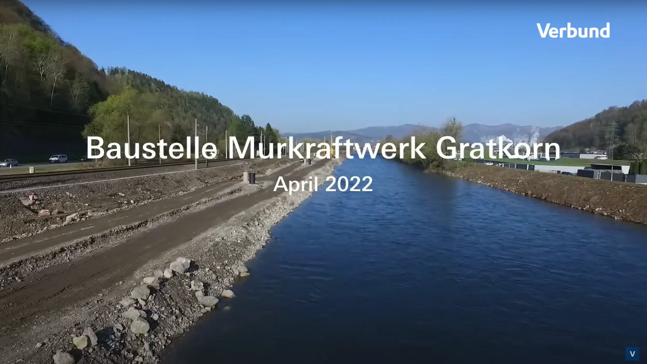 Luftaufnahme der Baustelle Murkraftwerk Gratkorn im April 2022. Das Bild zeigt einen Abschnitt des Flusses Mur mit klarem blauen Wasser und die umliegende Baustelle mit Schotterwegen und Baumaschinen bei sonnigem Wetter.